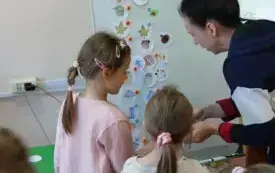dwie dziewczynki i pani stoją przed tablicą na kt&oacute;rej przyczepione są okrągłe karteczki z symbolami jesiennymi