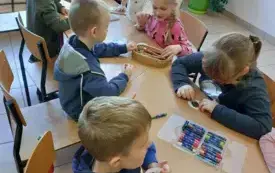 dzieci siedzą przy stoliku przed nimi leżą kredeki i papierowe k&oacute;łka na kt&oacute;rych dzieci rysują