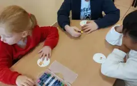 dziewczynka i dw&oacute;ch chłopc&oacute;w siedzą przy stoliku przed nimi leżą kredeki i papierowe k&oacute;łka na kt&oacute;rych dzieci rysują