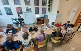 dzieci siedzą przy długim stole na kt&oacute;rym znajdują się kredki i k&oacute;łka z papieru pani siedzi przy tablicy na kt&oacute;rej przyczepione są emblematy z jesiennymi symbolami