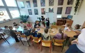 dzieci siedzą przy długim stole na kt&oacute;rym znajdują się kredki i k&oacute;łka z papieru pani siedzi przy tablicy na kt&oacute;rej przyczepione są emblematy z jesiennymi symbolami