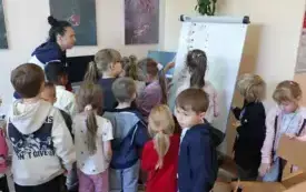 grupa dzieci stoi przed tablicą na kt&oacute;rej przyczepione są emblematy z jesiennymi symbolami wśr&oacute;d nich pani