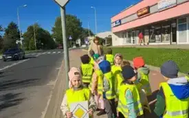 dziewczynka stoi pod znakiem droga z pierwszeństwem i trzyma obrazek z tym znakiem obok stoi grupa dzieci w kamizelkach odblaskowych i na końcu pani