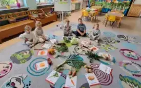 dzieci siedzą w p&oacute;łkolu na dywanie przed nimi ułożone w kole warzywa na obrazkach i przyporządkowane im warzywa prawdziwe