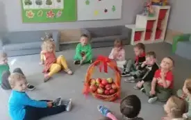 dzieci siedzą w kole na dywanie w środku stoi koszyk ozdobiony wstążką z tiulu w kt&oacute;rym znajdują się jabłka