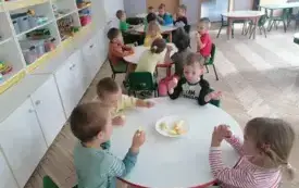 dzieci siedzą przy okrągłych stolikach i jedzą pokrojone jabłka z tależyka