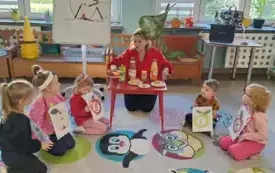 pani w przebraniu jabłka w czerwonej bluzie i opasce z listkiem siedzi za czerwonym stolikiem na kt&oacute;rym leżą produkty zrobione z jabłek przed stolikiem siedzą dzieci trzymające kartki z literami na szablonach jabłek