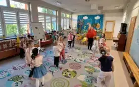 dzieci stoją w parach na sali trzymają literki kartki z literami w szablonie jabłka z tyłu stoi pani w czerwonej bluzie i opasce z listkiem widać tablićę na kt&oacute;rej wisi plakat z budową jabłka