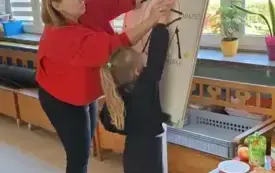 pani w przebraniu jabłka w czerwonej bluzie z opaską w kształcie liścia stoi przy tablicy wraz z dziewczynką na tablicy obrazek przedstawiający budowę jabłka