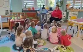 pani w przebraniu jabłka w czerwonej bluzie z opaska z liściem siedzi przed laptopem dzieci siedzą na dywanie patrzą na to co przedstawione jest na ekranie na ławce leżą rzeczy związane z jabłkami lub zrobione z jabłek