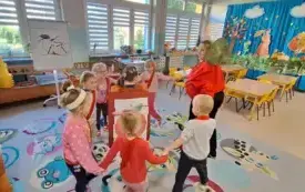 dzieci w małym k&oacute;łku tańczą na sali z nimi pani nauczycielka przebrana za jabłko w czerwonej bluzie i opasce z liściem w środku dziewczynka z plakatem czerwonego jabłka złożonego z element&oacute;w