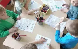 dzieci siedzą przy stoliku na kt&oacute;rym stoją kredki i leżą karty pracy