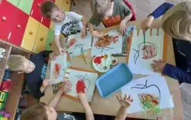 dzieci siedzą przy stoliku na kt&oacute;rym leżą farby dzieci malują folię bąbelkową na r&oacute;żne kolory pod folią leżą szablony jabłek