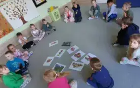 dzieci siedzą w kole na dywanie w środku leżą r&oacute;żne obrazki przedstawiające jabłka