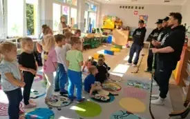 grupa dzieci z kt&oacute;rej część dzieci stoi część siedzi na dywanie przed nimi stoi czterech pan&oacute;w jeden z nich trzyma kij do hokeja