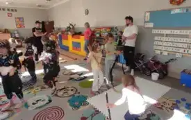 dzieci ubrane w kaski oraz ochraniacze oraz dzieci z kijami hokejowymi stoją na sali wśr&oacute;d nich panowie i pani 