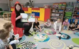 dzieci siedzą w kole na dywanie wśr&oacute;d nich na krzesełku siedzi pani w stroju czerwonego kapturka i trzyma otwartą książkę kt&oacute;rą pokazuje dzieciom