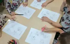 dzieci siedzą przy stoliku na kt&oacute;rym leżą przed nimi kolorowanki i kredki