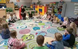 dzieci siedzą w kole na dywanie wśr&oacute;d nich na krzesełku siedzi pani w stroju czerwonego kapturka i trzyma książkę