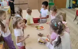 dzieci jedzą pokrojone warzywa i owoce kt&oacute;re stoją na talerzach na stoliku