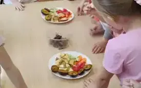 dzieci jedzą pokrojone warzywa i owoce kt&oacute;re stoją na talerzach na stoliku
