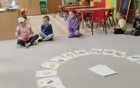 dzieci siedzą w kole w środku leżą obrazki warzyw i owoc&oacute; dziewczynka z zakrytymi oczami rozpoznaje za pomocą zmysł&oacute;w dotyku zapachu co trzyma w dłoni