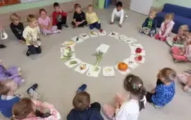 dzieci siedzą w kole na dywanie wewnątrz koła obrazki warzyw i owoc&oacute;w a na nich prawdziwe owoce i warzywa