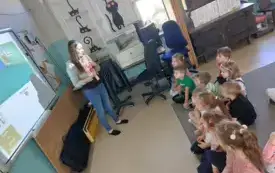 dzieci siedzą na dywanie przed tablicą multimedialną na kt&oacute;rej widać obrazki przy tablicy stoi pani i m&oacute;wi do dzieci pokazując im naklejki kt&oacute;re trzyma 