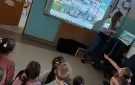 dzieci siedzą na dywanie przed tablicą multimedialną na kt&oacute;rej widać obrazki przy tablicy stoi pani i m&oacute;wi do dzieci