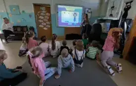 dzieci siedzą na dywanie przed tablicą multimedialną na kt&oacute;rej widać obrazki przy tablicy stoi pani i m&oacute;wi do dzieci