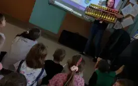dzieci siedzą na dywanie przed tablicą multimedialną na kt&oacute;rej widać obrazek 2 przy tablicy stoi pani i trzyma duże liczydło