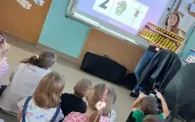 dzieci siedzą na dywanie przed tablicą multimedialną na kt&oacute;rej widać obrazek 2 przy tablicy stoi pani i trzyma duże liczydło