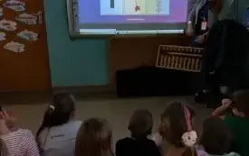 dzieci siedzą na dywanie przed tablicą multimedialną na kt&oacute;rej widać obrazek 1 przy tablicy stoi pani 