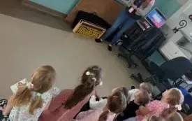 dzieci siedzą na dywanie przed tablicą multimedialną na kt&oacute;rej widać obrazki przy tablicy stoi pani i m&oacute;wi do dzieci