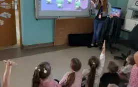 dzieci siedzą na dywanie zwr&oacute;cone w stronę tablicy multimedialnej przy kt&oacute;rej stoi pani na tablicy widać obrazki żabek