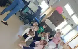 dzieci siedzą na dywanie przed nimi stoi pani tyłem do biektywu