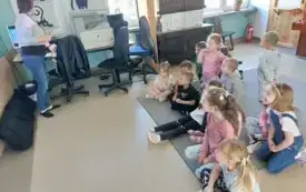 dzieci siedzą na dywanie przed nimi stoi pani przy komputerze