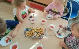 dzieci siedzą przy stoliku i jedzą sałatkę owocową na środku stołu stoi naczynie z sałatką