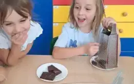 dwie dziewczynki przy stoliku jedna z nich trze czekoladę na tarce druga pochyla się do przodu przed nimi stoi naczycie z owocami i galaretką