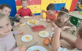 dzieci siedzą przy stoliku na kt&oacute;rym stoją talerzyki z owocami i naczynie z galaretką dzieci kroją owoce