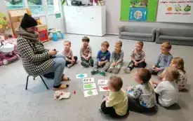 dzieci siedzą na dywanie w p&oacute;łkolu przed nimi leżą na dywanie obrazki z elementami garderoby pani siedzi na krzesełku w ubraniu zimowym