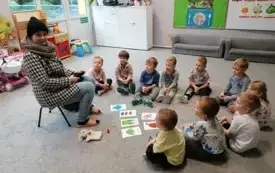 dzieci siedzą na dywanie w p&oacute;łkolu przed nimi leżą na dywanie obrazki z elementami garderoby pani siedzi na krzesełku w ubraniu zimowym