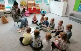 dzieci siedzą na dywanie w p&oacute;łkolu przed nimi leżą na dywanie obrazki z elementami garderoby pani siedzi na krzesełku w ubraniu zimowym