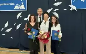 trzy elegancko ubrane panie oraz pan w garmiturze pozują do zdjęcia na tle dekoracji dotyczącej dnia edukacji narodowej panie trzymają kwiaty i dyplomy