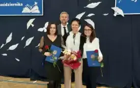 trzy elegancko ubrane panie oraz pan w garmiturze pozują do zdjęcia na tle dekoracji dotyczącej dnia edukacji narodowej panie trzymają kwiaty i dyplomy
