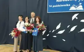 trzy elegancko ubrane panie oraz pan w garmiturze pozują do zdjęcia na tle dekoracji dotyczącej dnia edukacji narodowej panie trzymają kwiaty i dyplomy