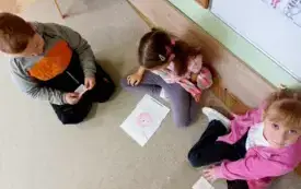 dwie dziewczynki i chłopiec siedzą na dywanie przed nimi ułożony obrazek kresk&oacute;wkowego wirusa