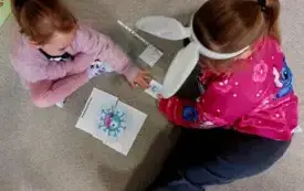 dwie dziewczynki siedzą na dywanie przed nimi ułożony obrazek kresk&oacute;wkowego wirusa