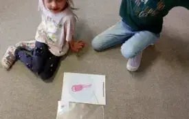 dwie dziewczynki siedzą na dywanie przed nimi ułożony obrazek kresk&oacute;wkowej bakterii