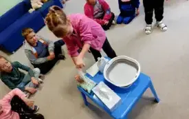 dziewczynka stoi przed niebieskim stolikiem na kt&oacute;rym stoi mydło w płynie, miska i leżą chusteczki dziewczynka wyciąga z paczki mokrą chusteczkę chusteczkę